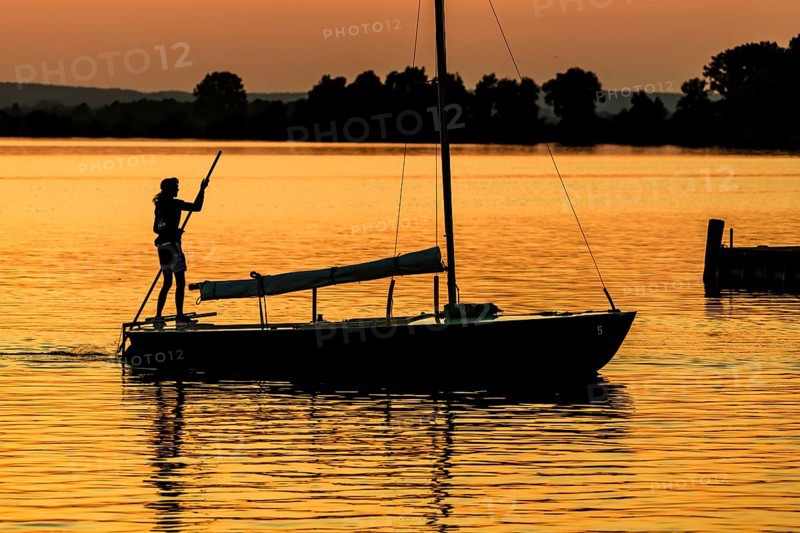 Sailing boat with person on calm Dümmer See brought into the harbor with a long stake at sunset, silhouette against orange sky, Lembruch, Dümmer See, Lower Saxony, Germany