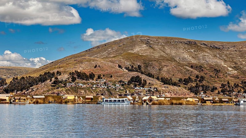 View of a lakeside village with hills in the background and clouds in the sky, The floating reed islands of the Uros in Lake Titicaca in Peru