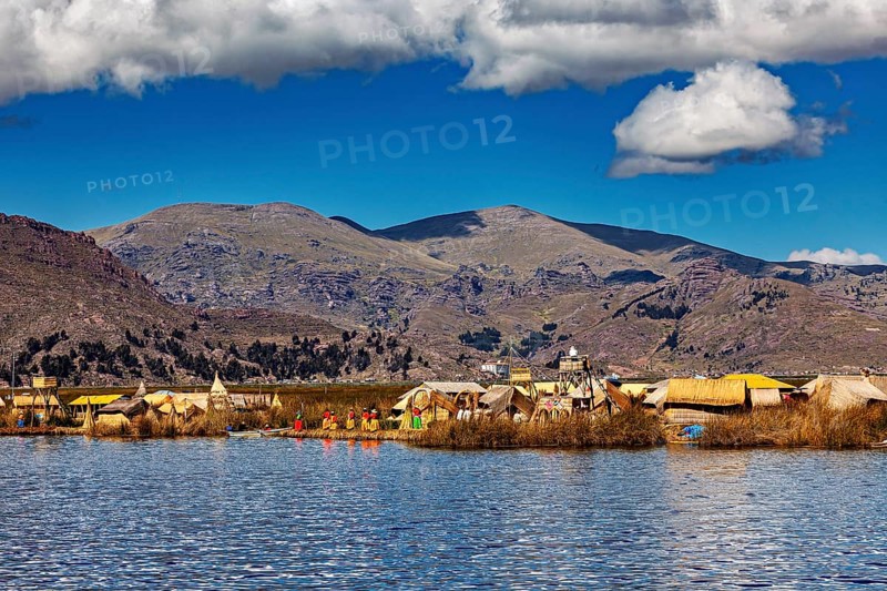 Scene with traditional huts on a lake against a mountain backdrop under blue skies and clouds, The floating reed islands of Uros in Lake Titicaca in Peru