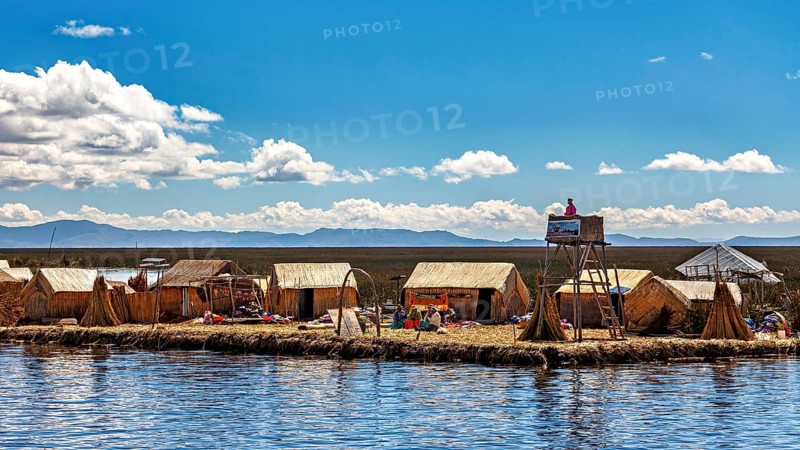 Reed huts and people on an island under a partly cloudy sky with a wide landscape, The floating reed islands of the Uros in Lake Titicaca in Peru