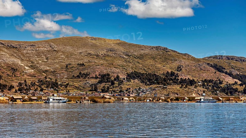 A calm lake in front of a mountainous landscape with boats and a village on the shore under a blue sky, The floating reed islands of the Uros in Lake Titicaca in Peru