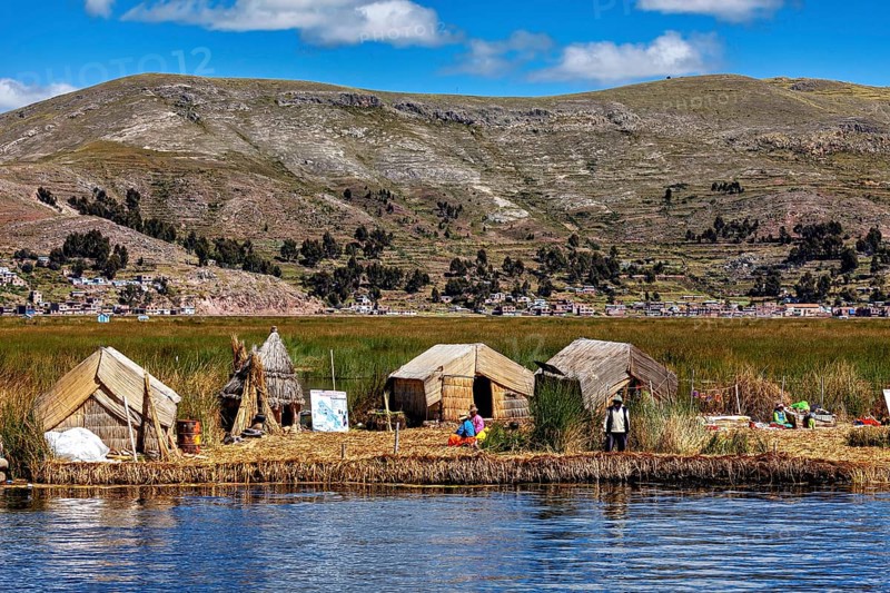 Huts on a reed island in front of a village and mountains, reflecting in the water, The floating reed islands of the Uros in Lake Titicaca in Peru