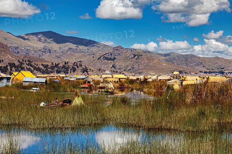 A rural area with huts, reeds and boats against a mountain backdrop and blue skies, The floating reed islands of the Uros in Lake Titicaca in Peru