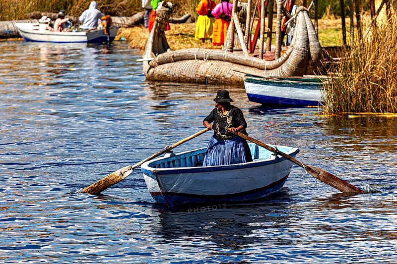 A woman in traditional dress rows a boat across a calm lake, the floating reed islands of the Uros in Lake Titicaca in Peru