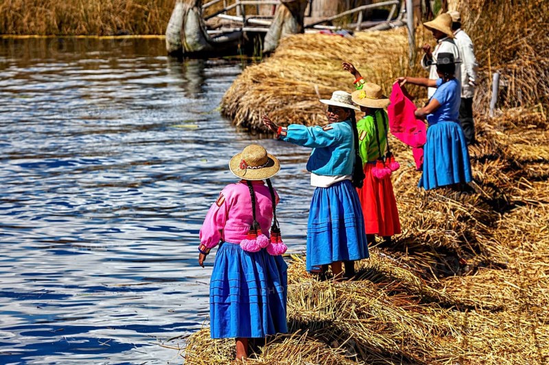 Women in colorful traditional clothing stand on the shore and look out at the water, the floating reed islands of the Uros in Lake Titicaca in Peru