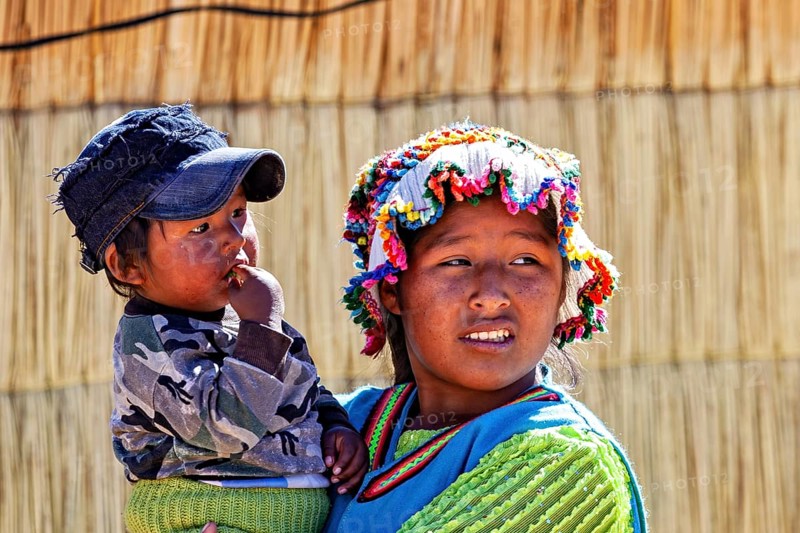 Woman wearing colorful hat carrying a child in her arms, both in traditional clothing, The floating reed islands of the Uros in Lake Titicaca in Peru