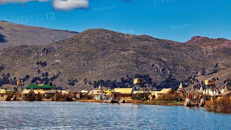 Huts on the shores of a lake in front of a mountainous landscape under a blue sky, The floating reed islands of the Uros in Lake Titicaca in Peru