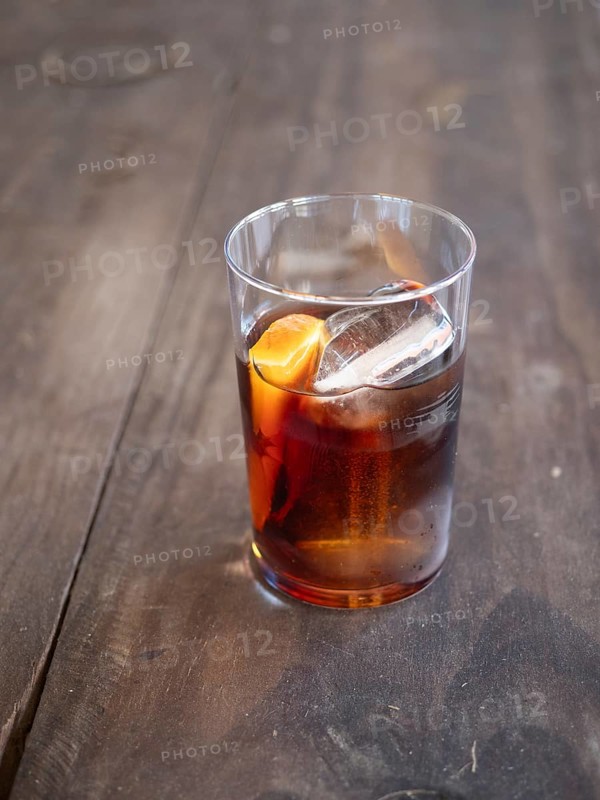 Glass of dark alcoholic cocktail with ice and orange zest on a rustic wooden table