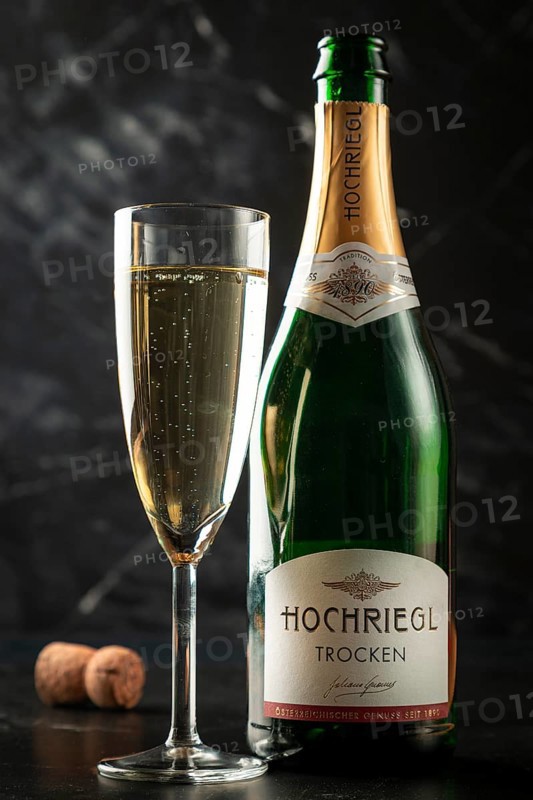 A champagne bottle next to a glass with sparkling contents, cork in the background