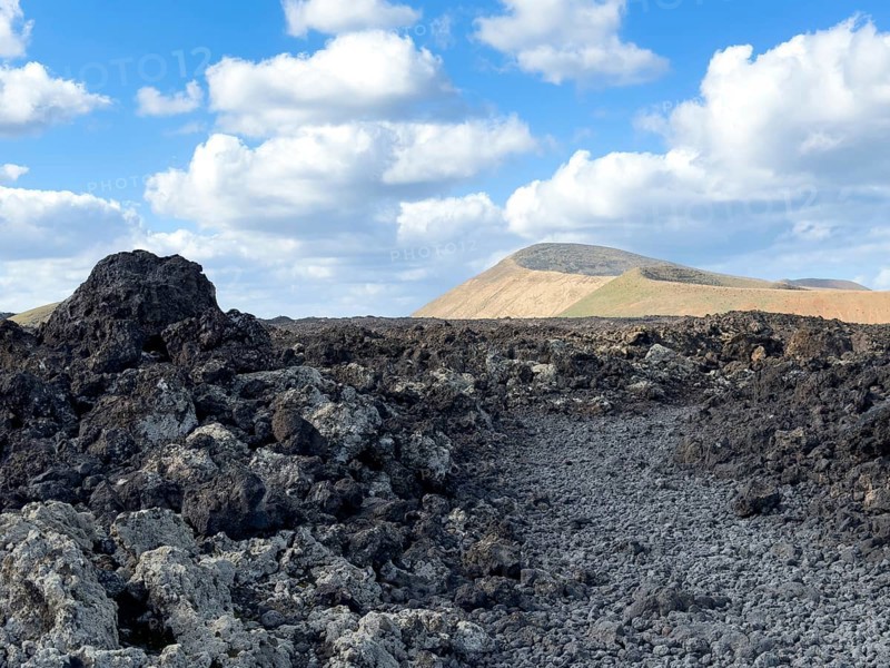 Hiking trail through lava rock to Caldera Blanca volcano against blue sky with white clouds, Mancha Blanca, Lanzarote, Canary Islands, Spain