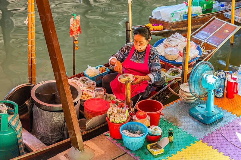 Elderly woman wearing mask offers food on her wooden boat, Damneon Saduak floating markets channel, near Bangkok, Ratchaburi district, Thailand