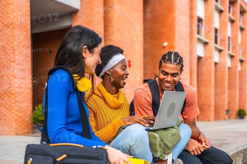 Group of diverse university students smiling and interacting outdoors, using a laptop, representing concepts of education, friendship, and collaborative learning on campus