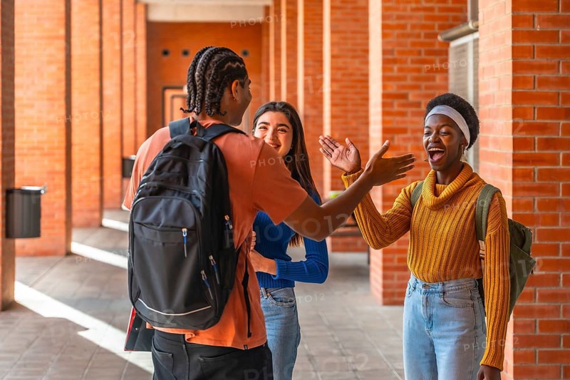 Diverse young students are happily greeting each other while walking along a brick wall corridor on a university campus, sharing a moment of friendship and connection