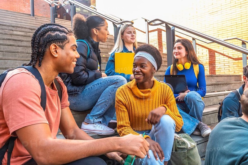 Diverse group of young students sitting on university campus steps, happy and smiling while discussing, representing education, friendship, and diversity in a college environment