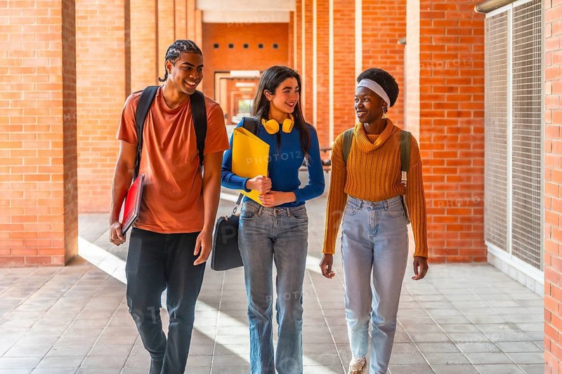 Diverse students walking through a university building on campus, carrying backpacks and notepads, smiling, and talking while enjoying their college education experience