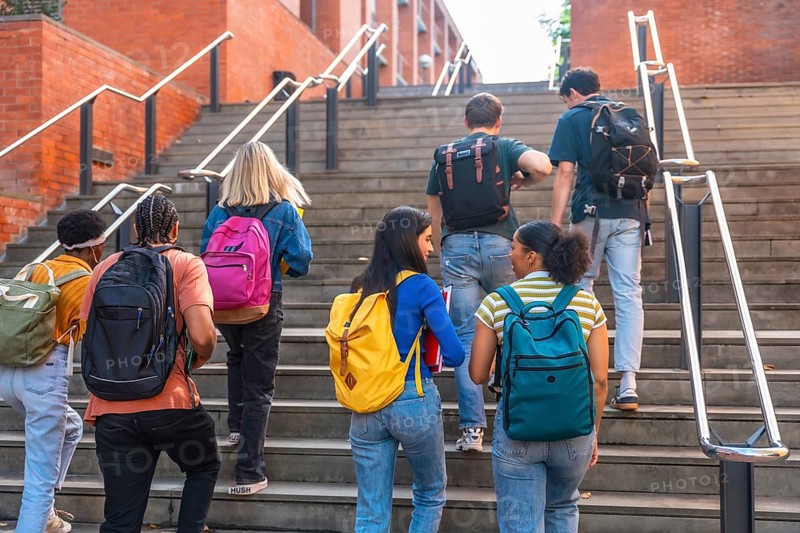 Group of young, multi ethnic university students with backpacks ascending outdoor stairs at a college campus, symbolizing education, learning, and the journey of academic life