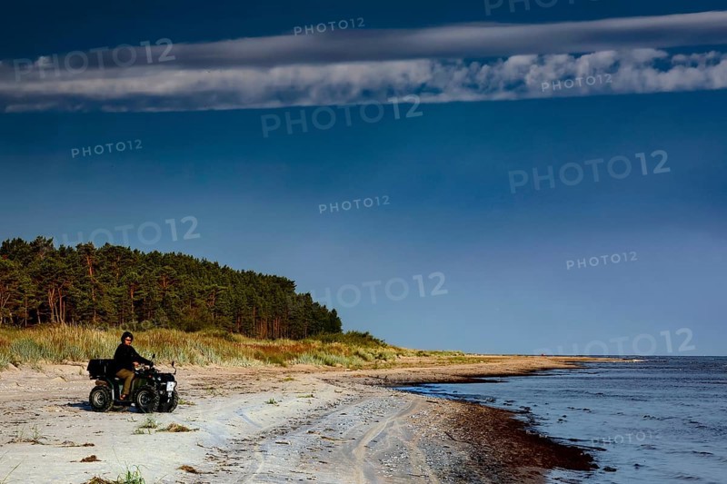 ATV rider rides along deserted coast with forest in background under clear sky, Kolka, Latvia