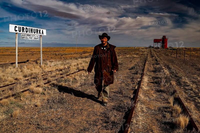 Man wearing cowboy clothes on abandoned tracks in front of station sign and water tower, Oodnadatta Track, Curdimurka, Australia