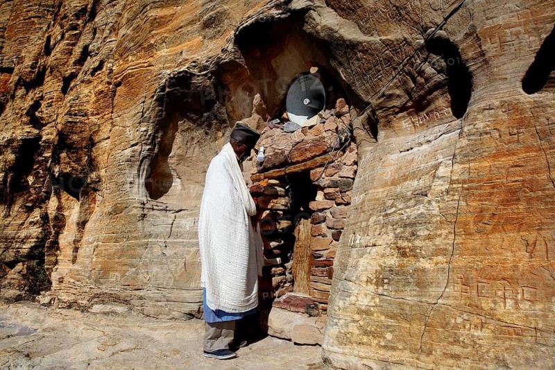 Priest stands in front of the rock church Abba Daniel Krokor in traditional clothing, zero