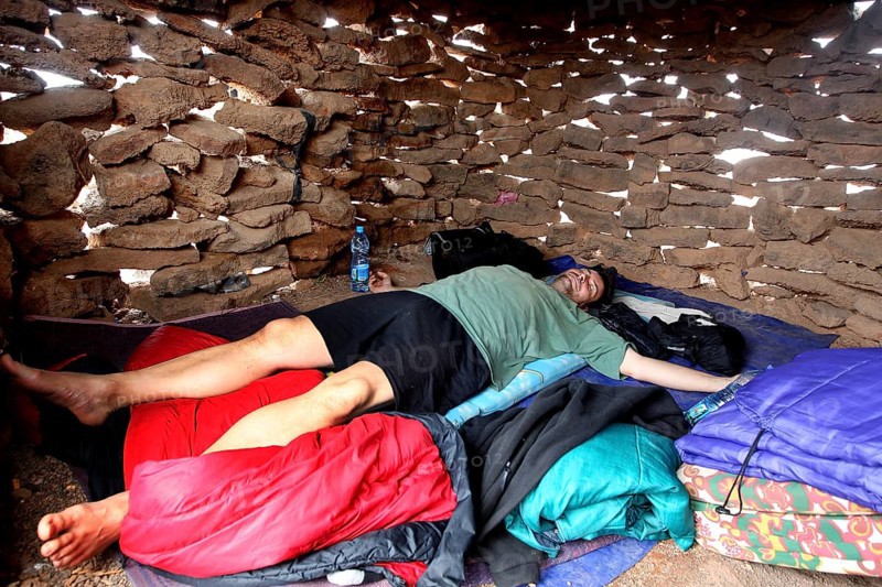 Person lying in a sleeping bag in a simple stone hut, zero