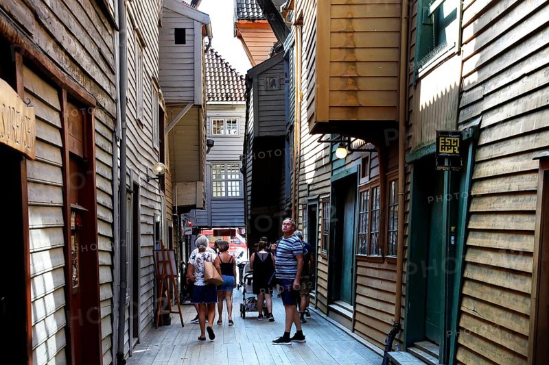 Narrow wooden house alley in Tyskebryggen with passers-by in sunshine, mountains, Hordaland, Norway