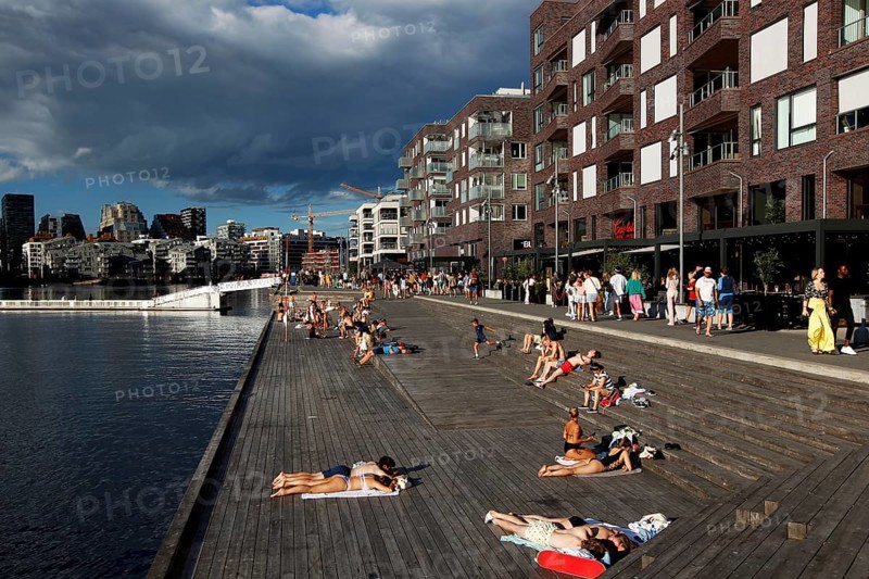 People relaxing on wooden walkway in the sun surrounded by new buildings, Oslo, Norway