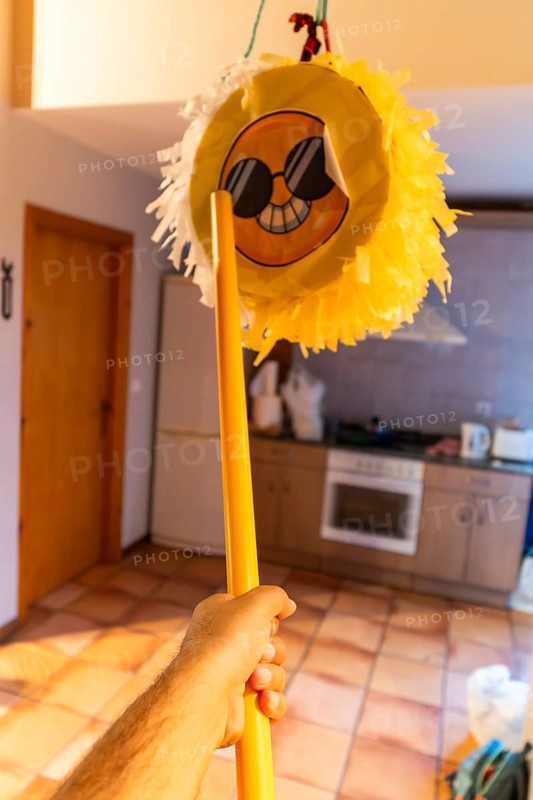 Hand holding yellow stick poised to strike a hanging emoji pinata in a home interior, playful birthday celebration scene with festive energy, candy and surprise imminent