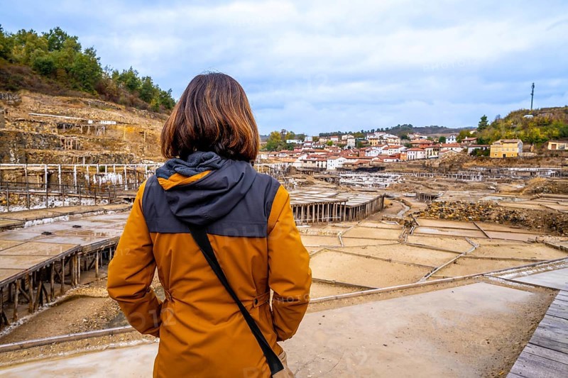 Woman tourist standing at anana salt valley, observing terraced evaporation ponds and traditional village houses against a rural basque landscape and cloudy autumn sky