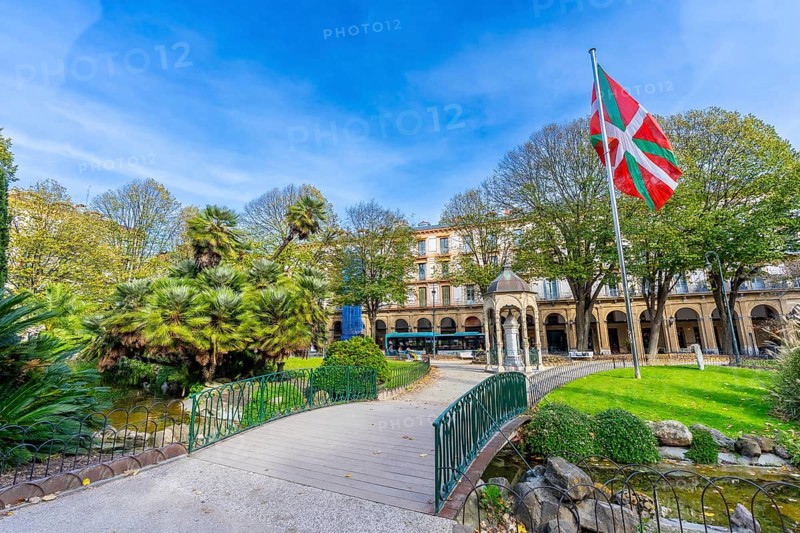 Gipuzkoa plaza park showing lush greenery, a pond with a small bridge, historic buildings, and the vibrant ikurrina flag flying proudly under a clear blue sky in san sebastian, basque country