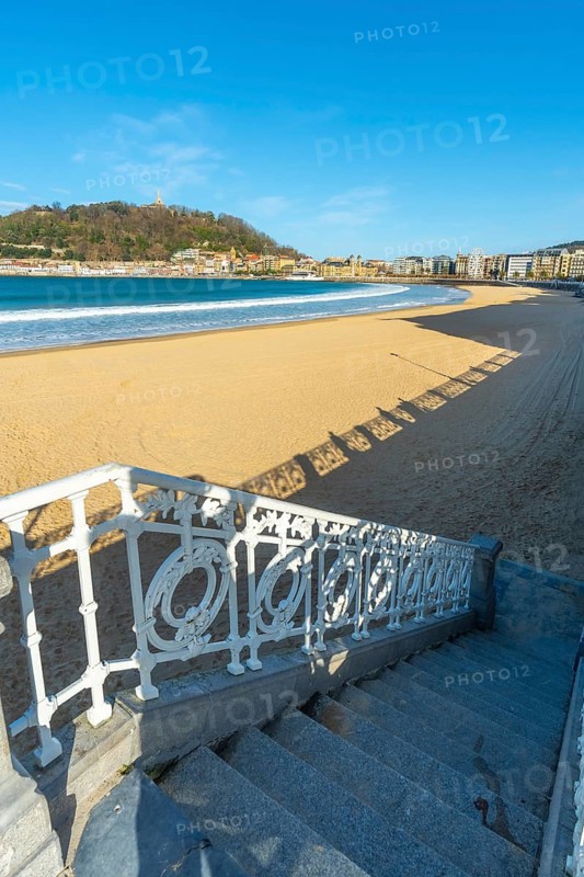 Scenic view looking down ornate white railings and stairs to the wide sandy beach of la concha bay, with the city and monte igueldo hill in the distance under a clear blue sky
