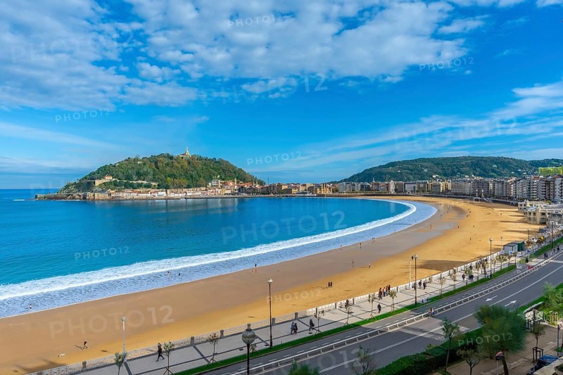 La concha beach curving along the bay of san sebastian city, with people walking on the sand and promenade, overlooking monte igueldo and the urban coastline under a bright blue sky