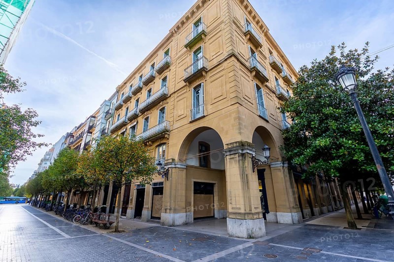 Gipuzkoa square in san sebastian, basque country, with historic facades, wrought iron balconies and stone archways lining a tree shaded pedestrian street under blue sky