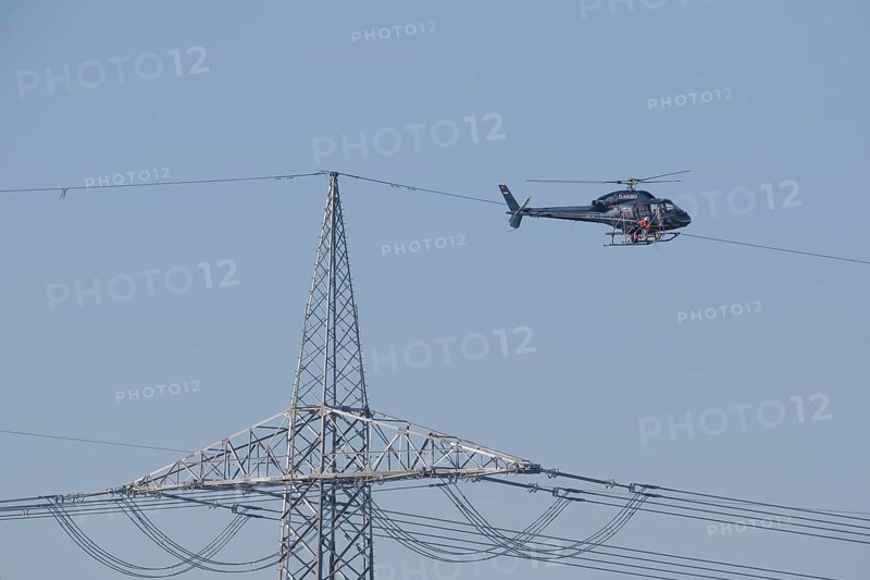 A helicopter carries out technical work on power lines next to a mast under a blue sky, an employee marks the top line of a new power line, Lemförde, Lower Saxony, Germany
