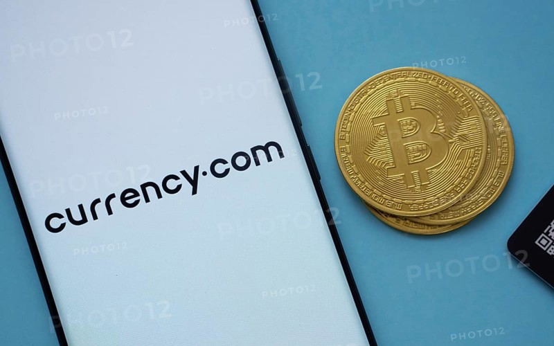 Dhaka, Bangladesh- 22 June 2025: Currency.com logo on smartphone screen near Bitcoin coin and QR code on blue background