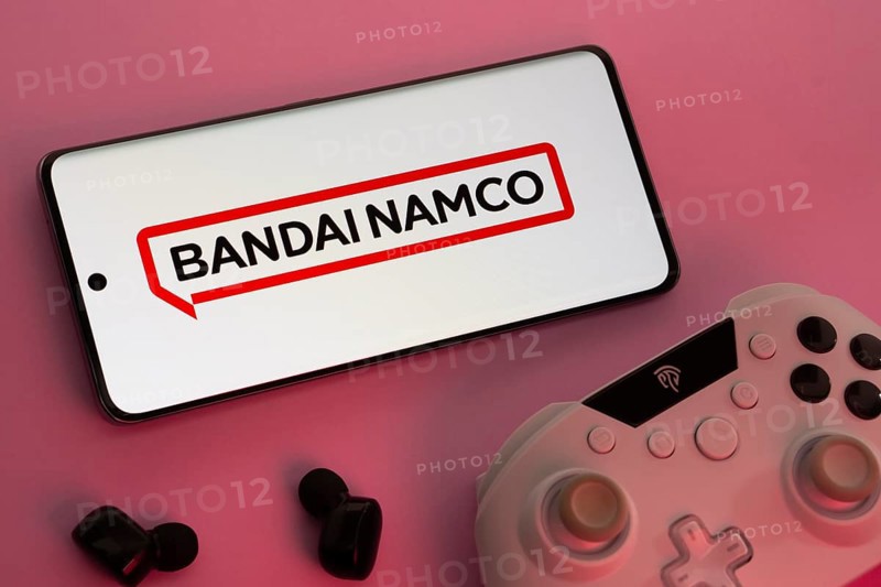 Dhaka, Bangladesh- 08 Apr 2025: A smartphone displaying the Bandai Namco Entertainment logo next to a gaming controller and earbuds on a pink surface