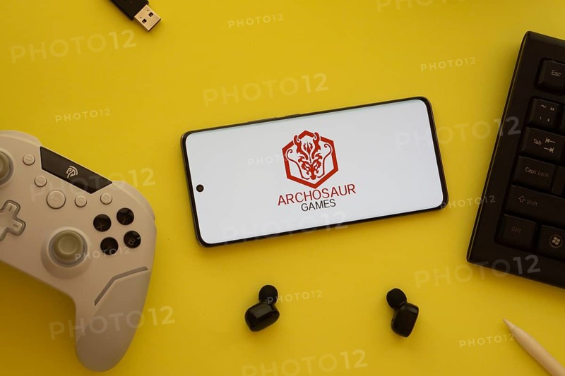 Dhaka, Bangladesh- 23 March 2025: Archosaur Games logo displayed on a smartphone surrounded by gaming accessories in an yellow light