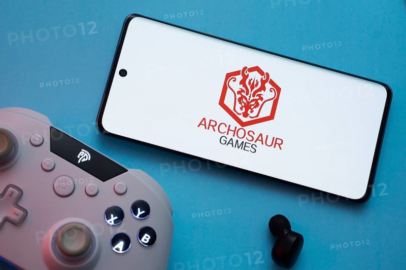 Dhaka, Bangladesh- 22 March 2025: Archosaur Games logo displayed on a smartphone surrounded by gaming accessories in an Blue background