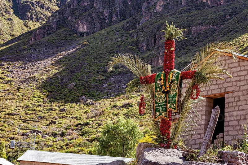 A scarecrow decorated with traditional costumes stands in a sunny mountain landscape next to a small building, cross in the landscape of the Colca Canyon of Peru