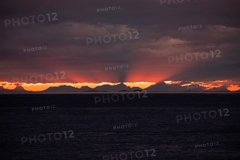 The Vestfjord in Nordland, Norway, offers a dramatic atmosphere near Bodø. Dark clouds are illuminated by the setting sun in shades of red and yellow. The rugged cliffs and mountains are reflected in the quiet fjord, which offers a breathtaking view of Lofoten