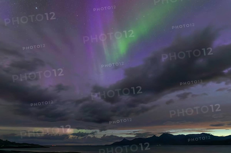 The Vestfjord in Nordland, Norway, has an enchanting atmosphere at night. Dark clouds and vivid aurora, polar lights dance over the fjord. The rugged cliffs and majestic mountains are reflected in the calm water and create a mystical backdrop