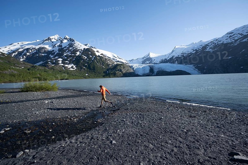 Young man jumping on a pebble beach on a glacial lake, Snowy Mountains and Portage Glacier on Portage Lake glacial lake, Chugach National Forest, Alaska, USA