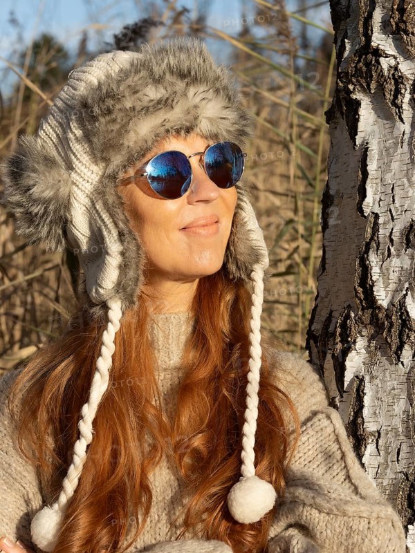 Slender pretty woman with fur hat and pompoms, red hair and warm winter colors between birch forest and reeds