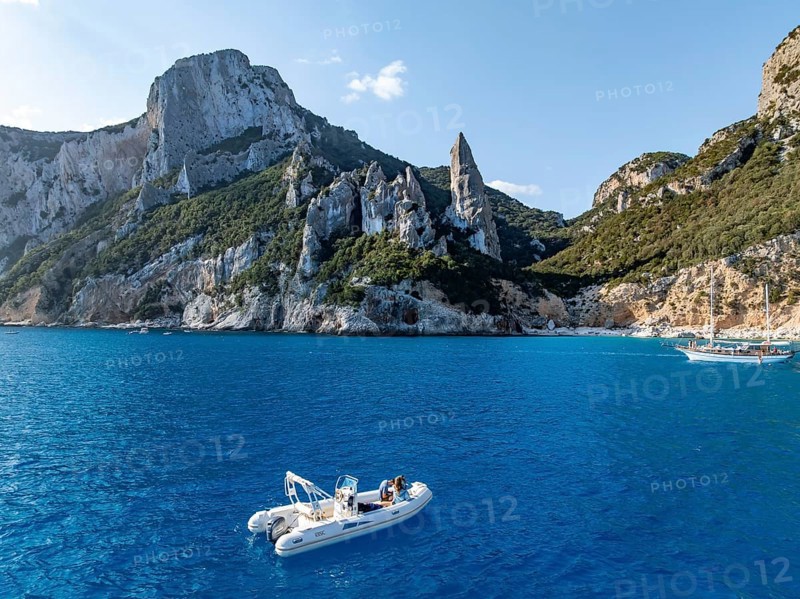 Motor boat off picturesque rocky coast, cliffs with L'Aguglia pinnacle, blue sea and Cala Goloritzé beach, aerial view, Golfo di Orosei, Baunei, Sardinia, Italy