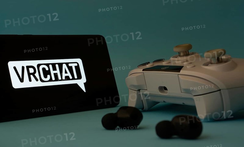 Dhaka, Bangladesh- 23 Mar 2025: A smartphone with a VRChat icon next to a controller and earbuds on a blue surface