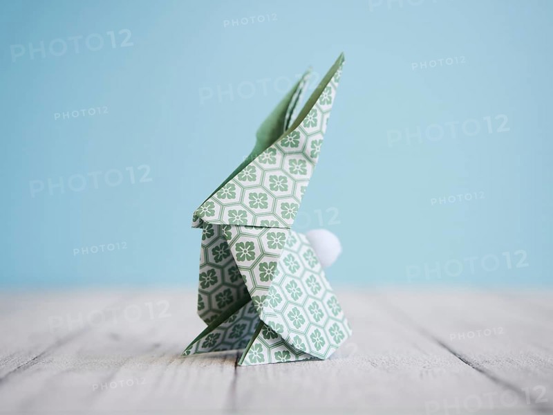 Green origami bunny standing on a white wooden table with a light blue background, representing easter and creativity