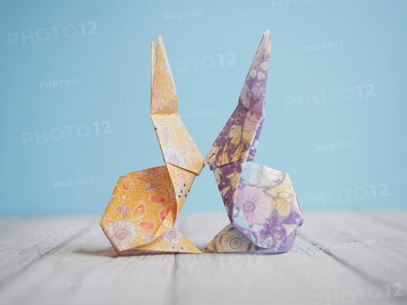 Two colorful origami bunnies are facing each other, touching noses, on a wooden surface with a light blue background