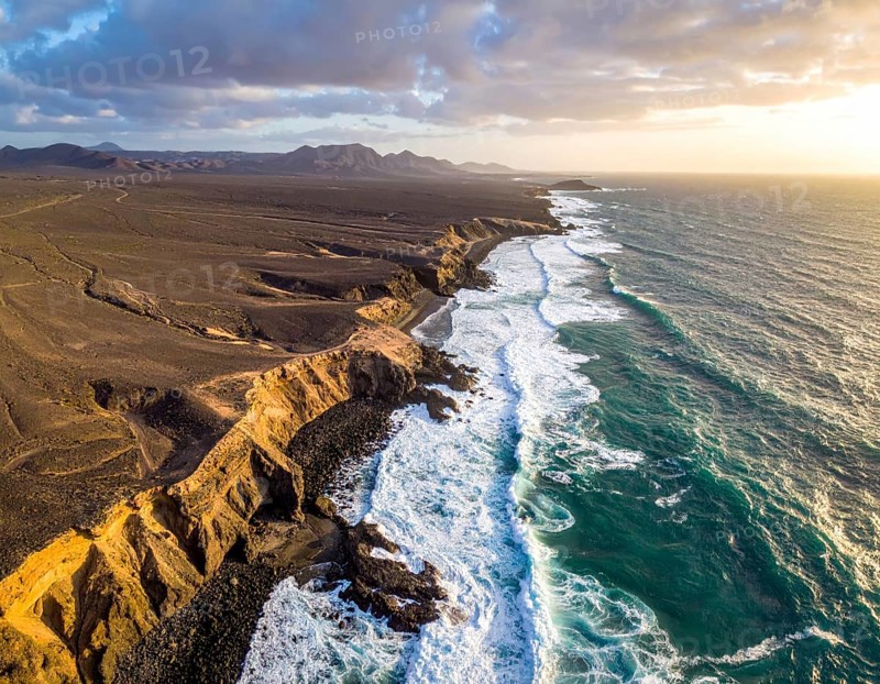Desert landscape converges with ocean along a rugged coastal area, shore break at cliffs, blue cloudy sky at sunset, dry landscape in an aerial drone view, AI generated