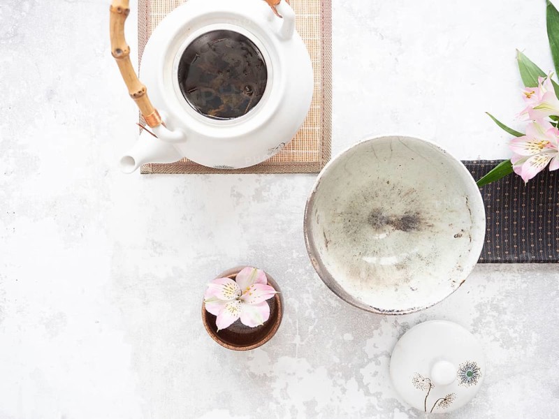 White teapot with bamboo handle, empty bowl, lid and pink flowers on light textured background are ready for tea ceremony