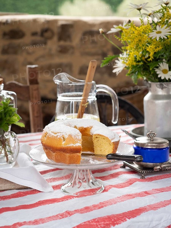 Slice of lemon cake with powdered sugar on a glass stand, pitcher of lemonade, flowers, and colorful tablecloth