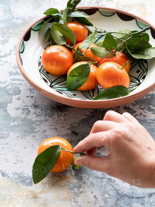 Fresh tangerines with leaves are being picked up from a ceramic bowl on a textured surface